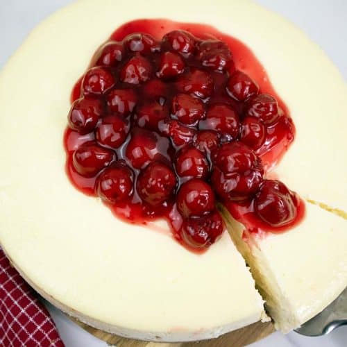 Vanilla cheesecake with a slice on a metal spatula and cherries on top.