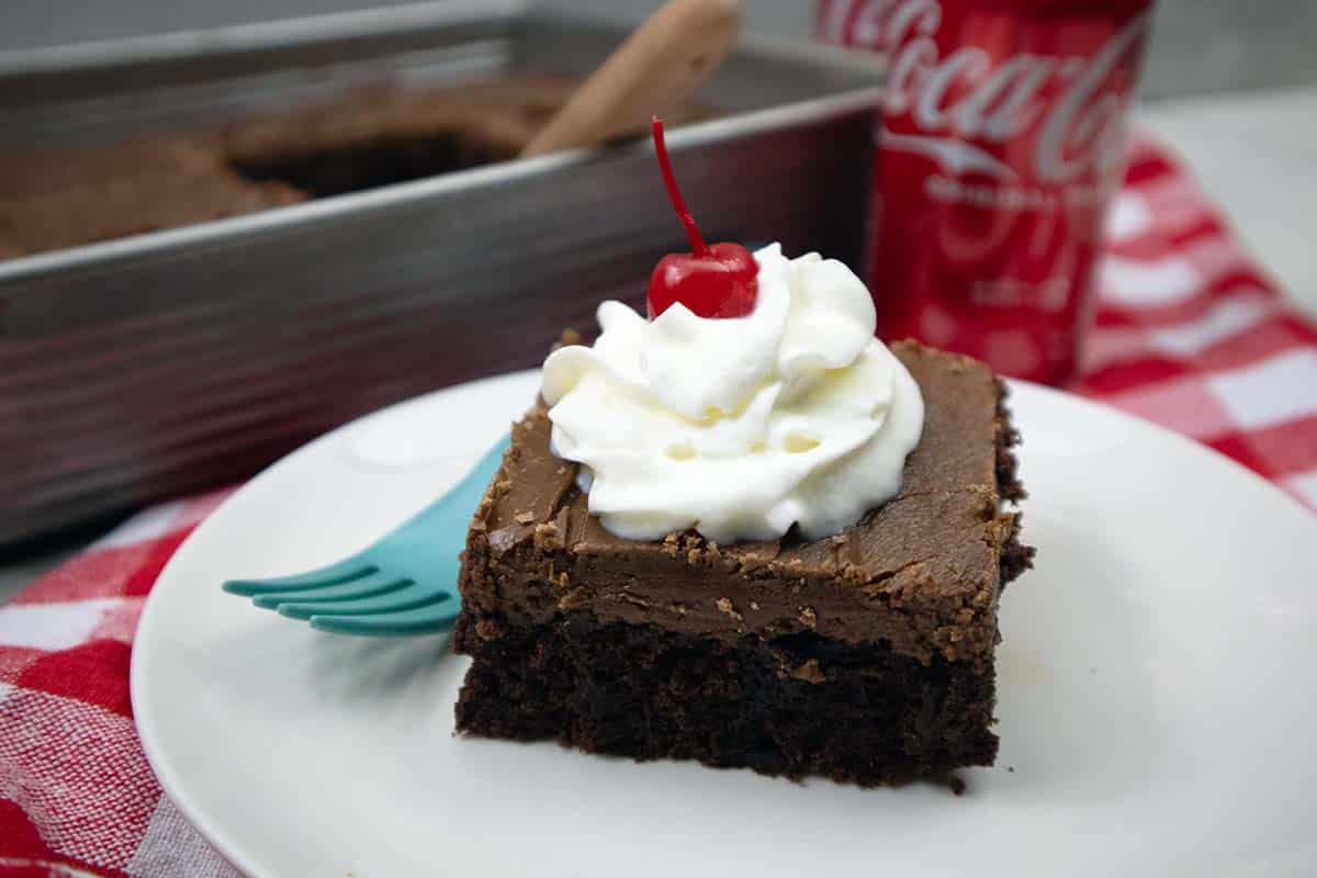 Coca Cola Cake slice on a white plate with cake in the background and a can of coca cola.
